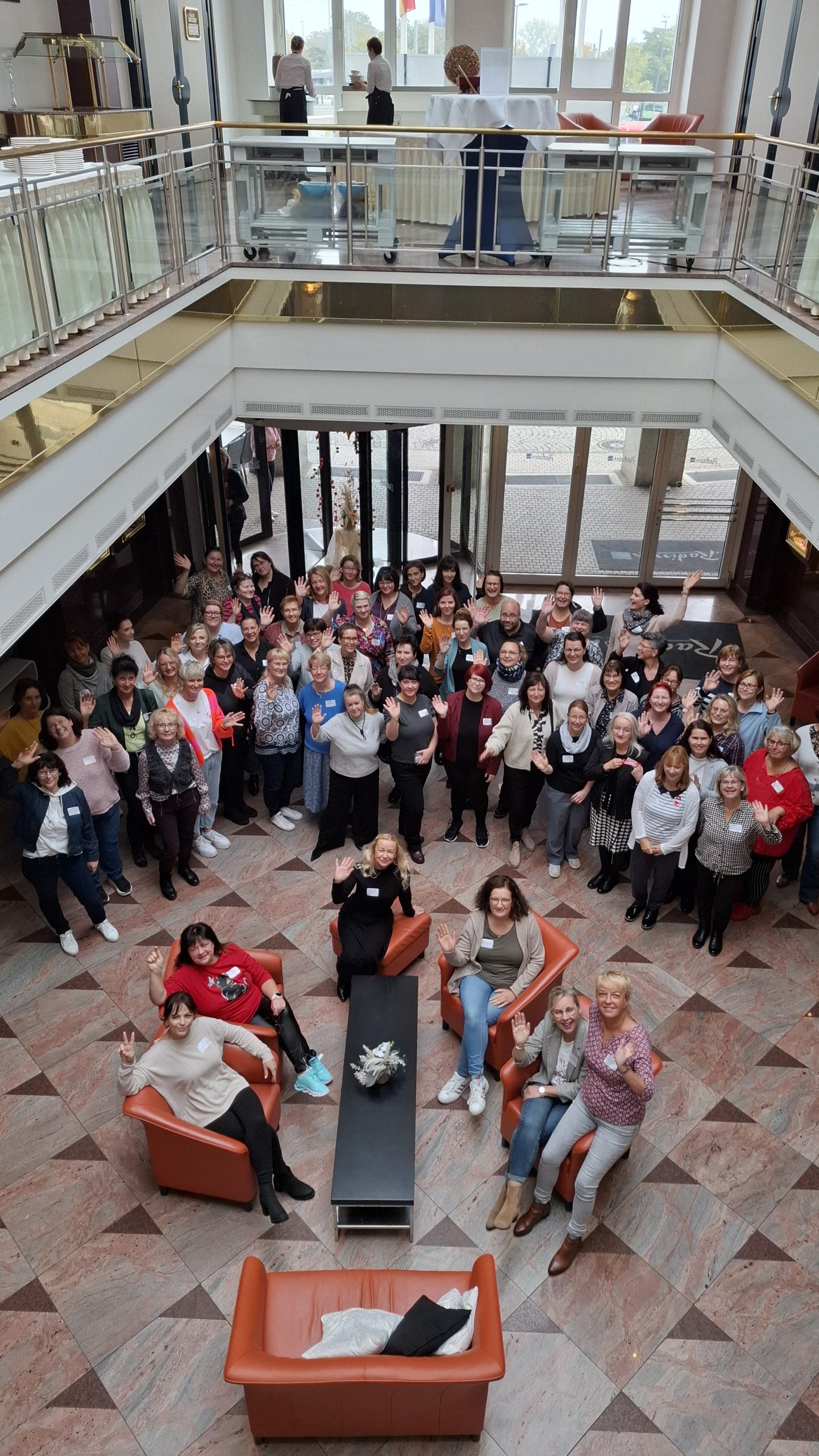Cottbus 20251015_105042 Viele Teilnehmende der Fachatung in Cottbus versammelten sich im Foyer für das Gruppefoto einige Personen sitzen auf Lounge Möbeln, die meisten anderen stehen im Halbkreis dahint, lächeln in die Kamera und Winken